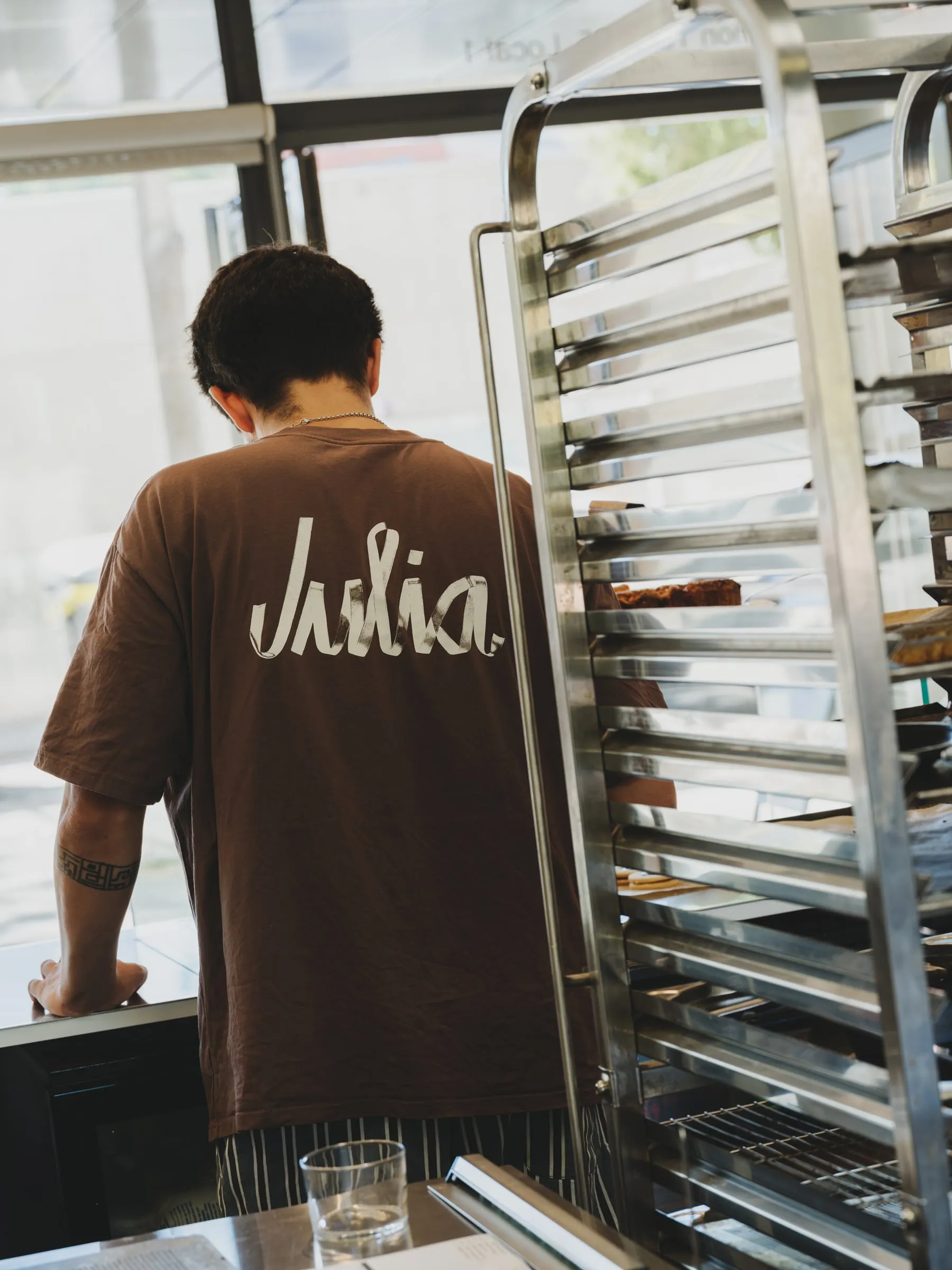 Camiseta marrón con el logotipo de Julia en blanco en la espalda, en la cocina de la cafetería junto a un carro de bandejas
