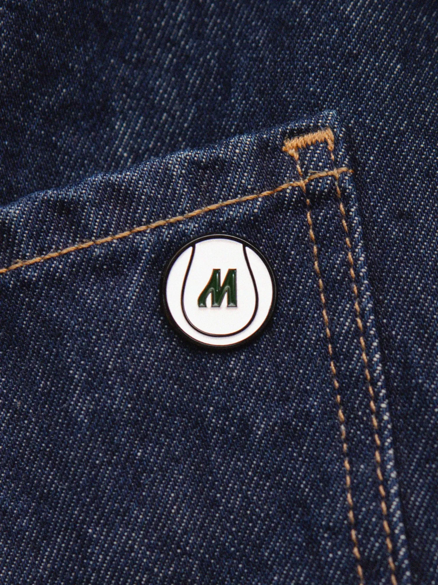 Chapa esmaltada con el emblema de Mostai, una pelota con una M, en el bolsillo de unos vaqueros