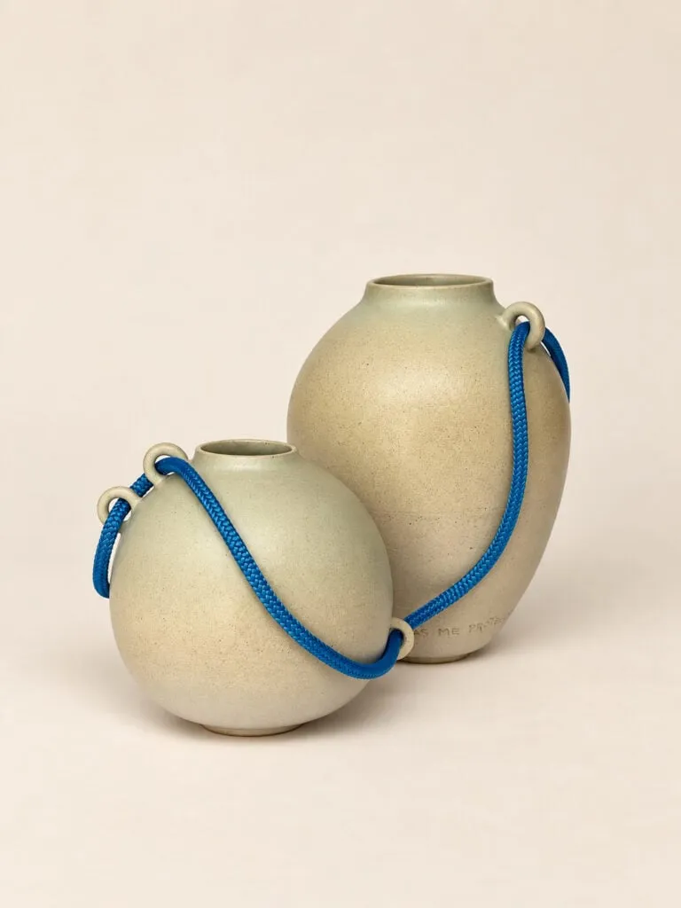 Dos vasijas de gres de distinto tamaño, unidas por un cordón azul que pasa por sus asas cerámicas, sobre fondo beige