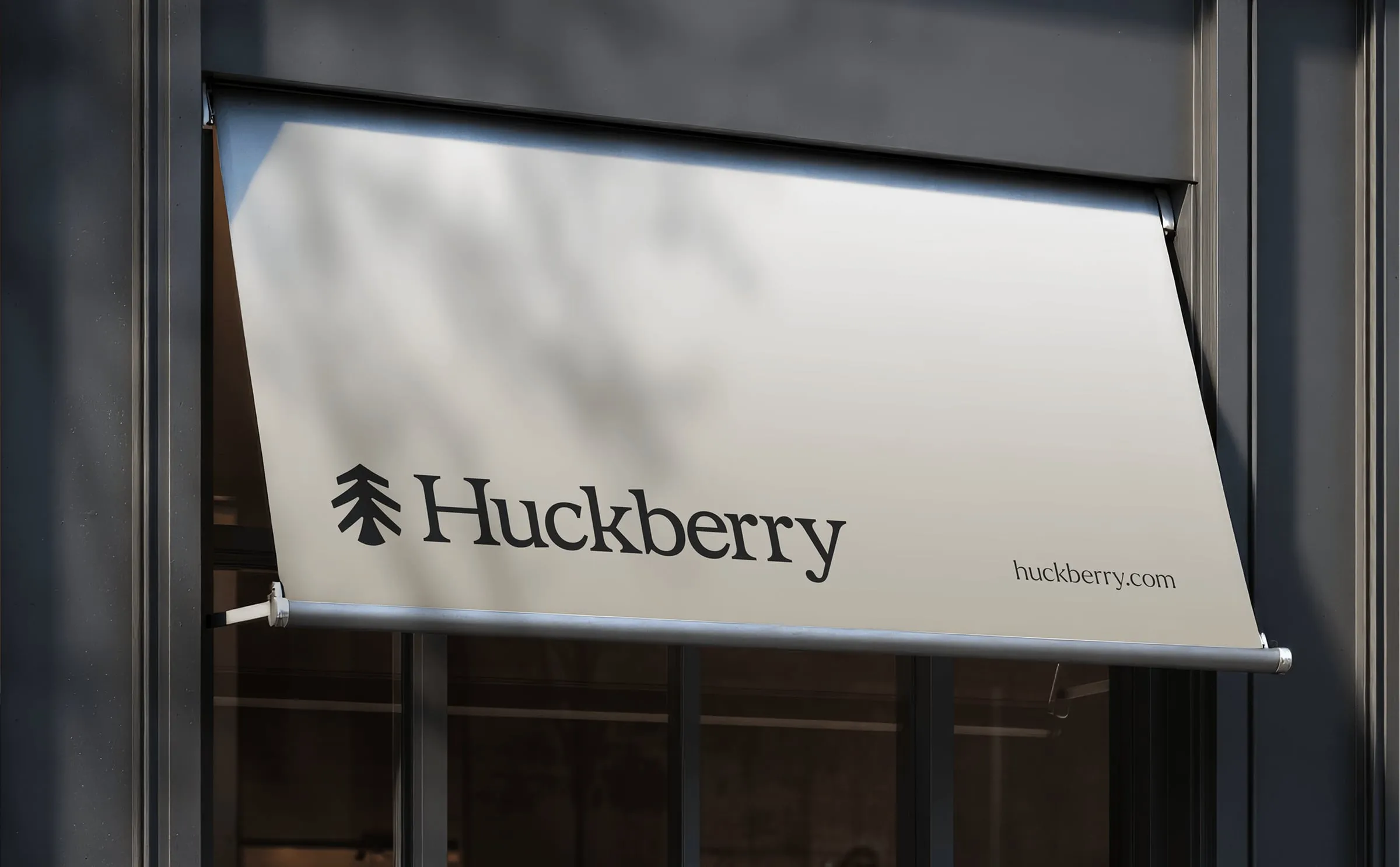 Toldo gris claro sobre el escaparate de una tienda con el logotipo de Huckberry, un pino estilizado junto al nombre, y la dirección huckberry.com