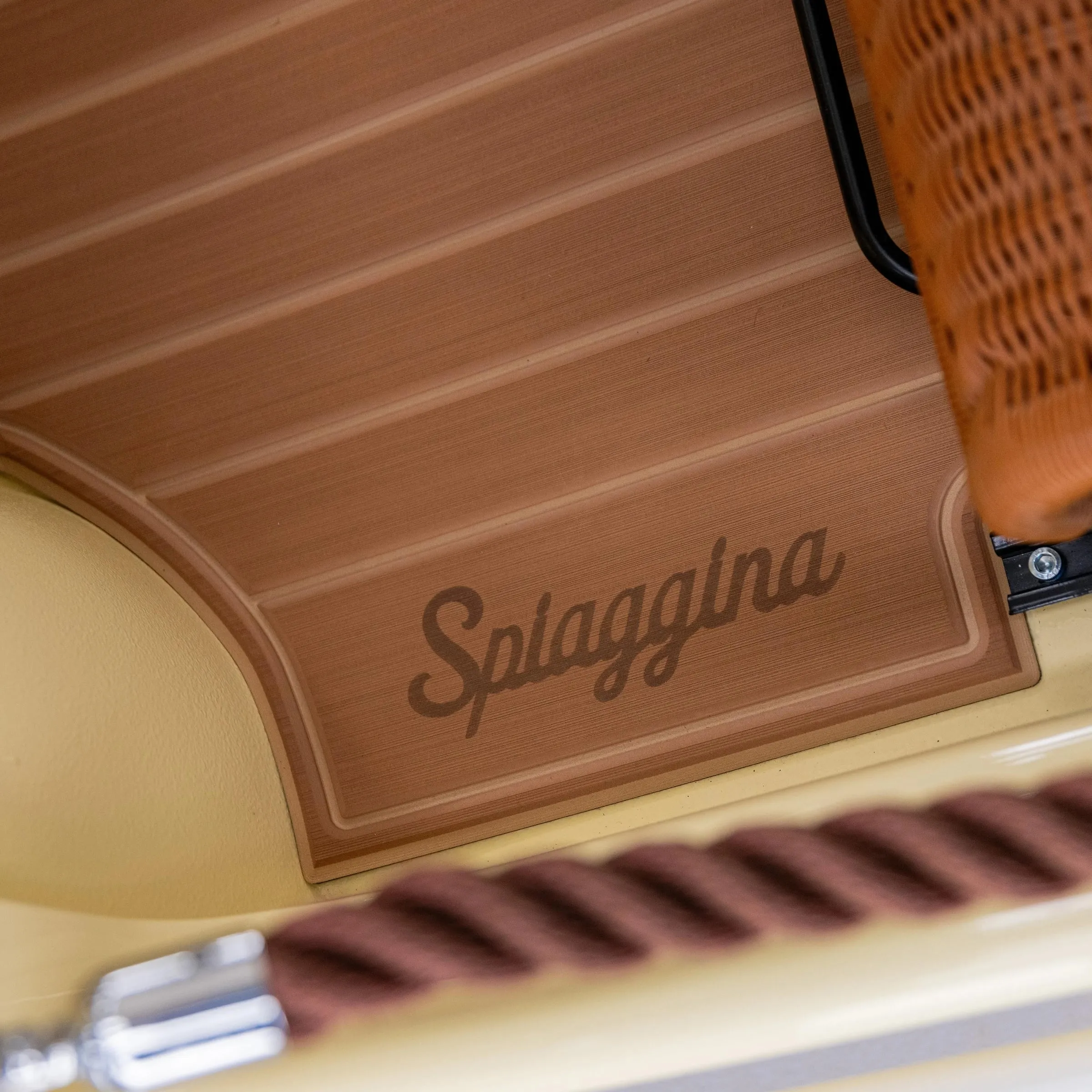 Suelo de imitación de madera con la palabra «Spiaggina» grabada, junto a un asiento de rattán y una cuerda gruesa trenzada de color burdeos