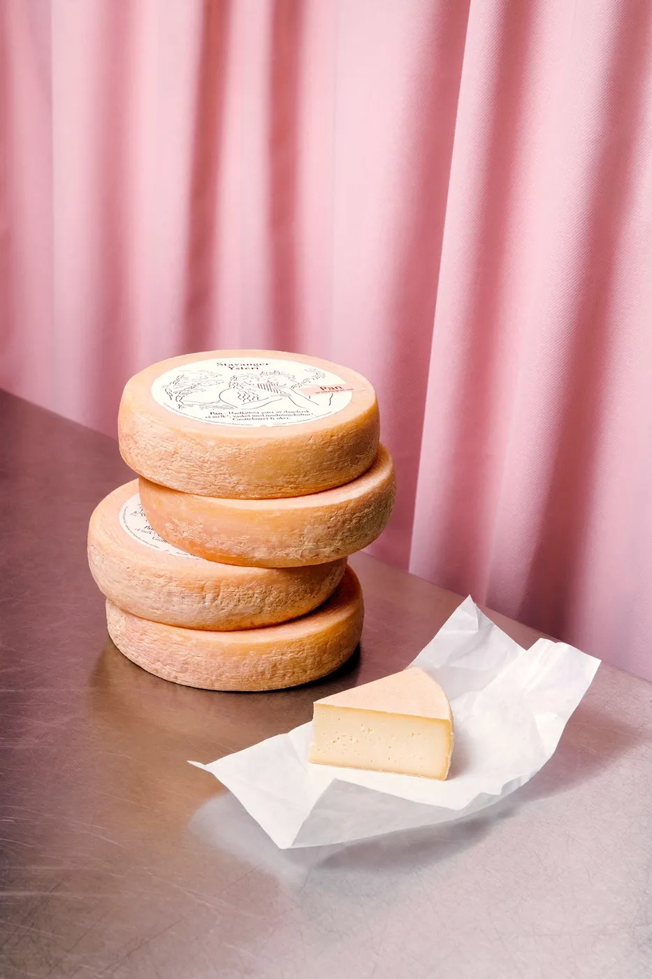 Pila de ruedas de queso rosado con una cuña sobre papel encerado arrugado, frente a una cortina rosa