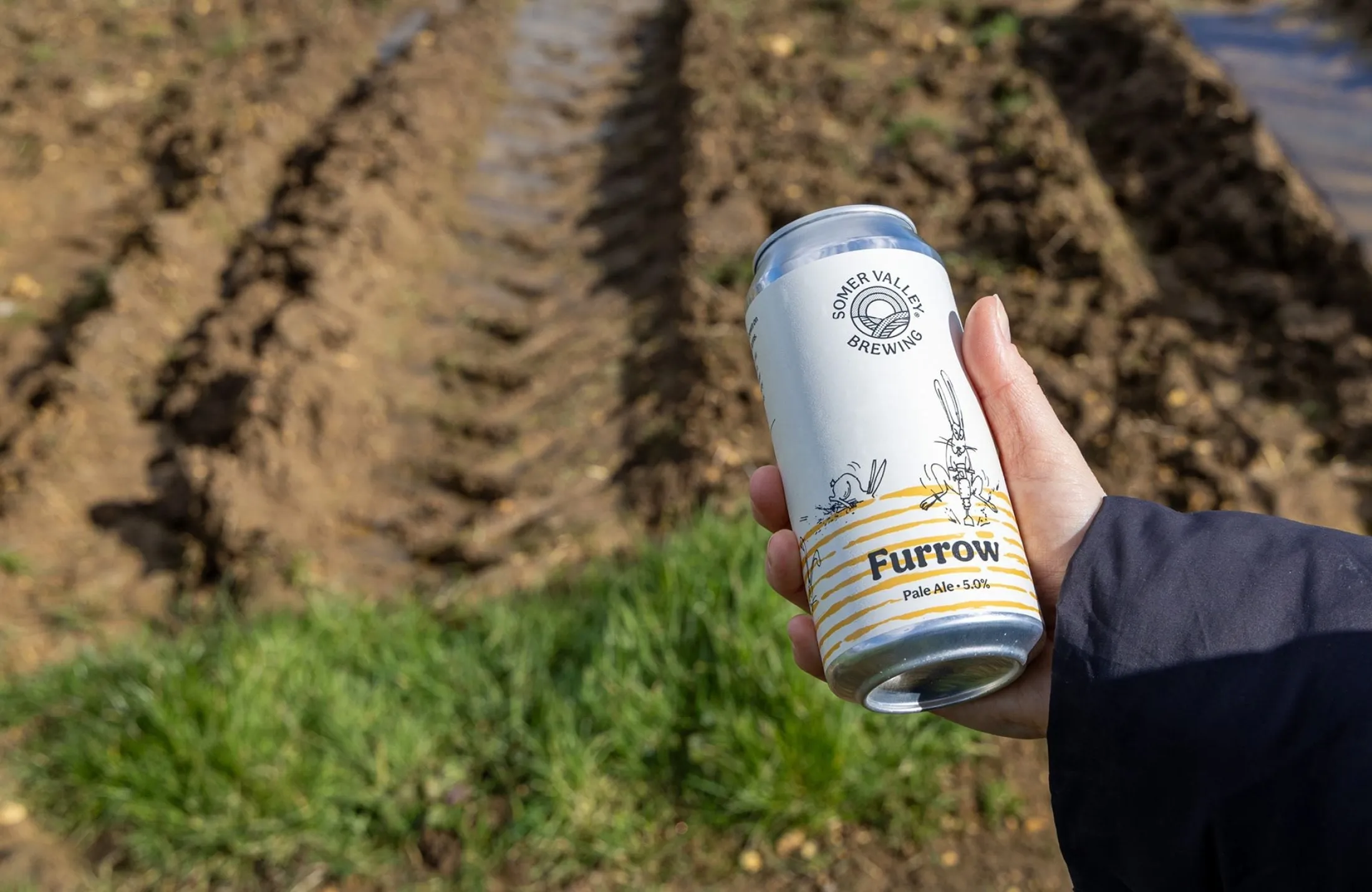 Mano sosteniendo una lata Furrow sobre una tierra labrada con surcos