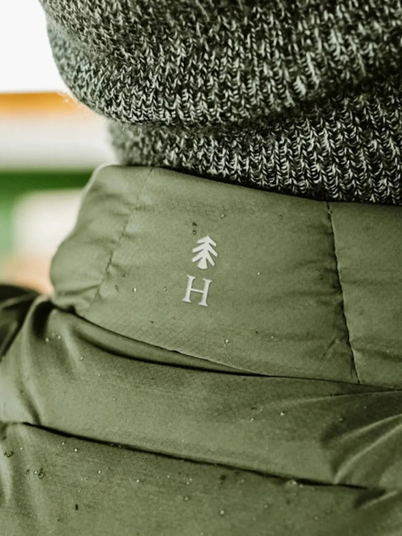 Primer plano del cuello de una chaqueta acolchada verde oliva con el pino de Huckberry y una H mayúscula en blanco, bajo un gorro de lana gris