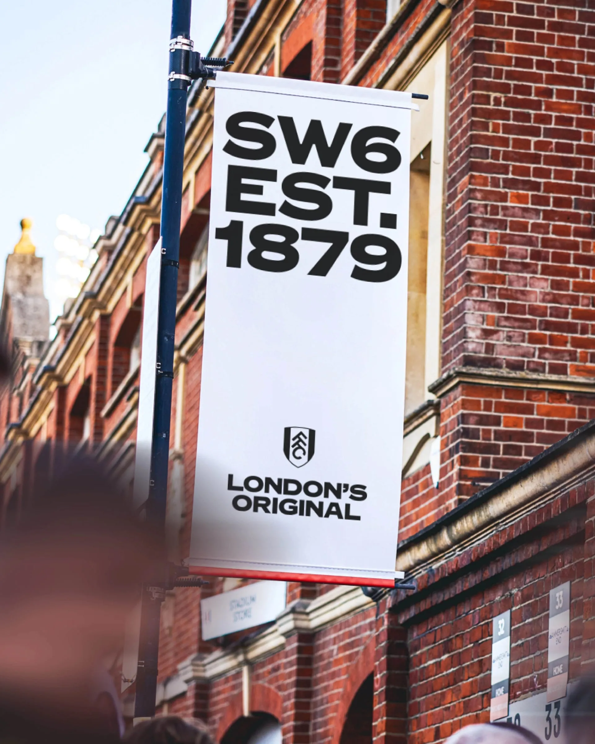 Estandarte blanco colgado de una farola ante la fachada de ladrillo del estadio, con «SW6 EST. 1879», el escudo y «London’s Original»