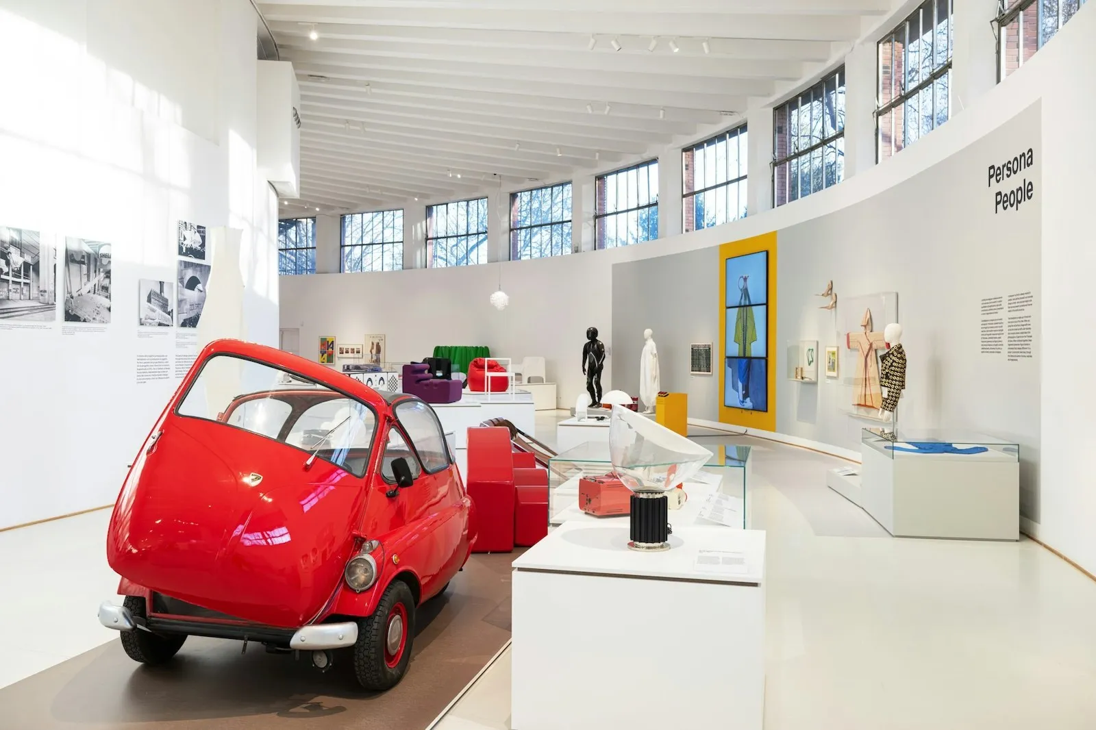 Sala curva con un microcoche rojo en primer plano, vitrinas blancas, una lámpara y una pantalla vertical sobre fondo amarillo