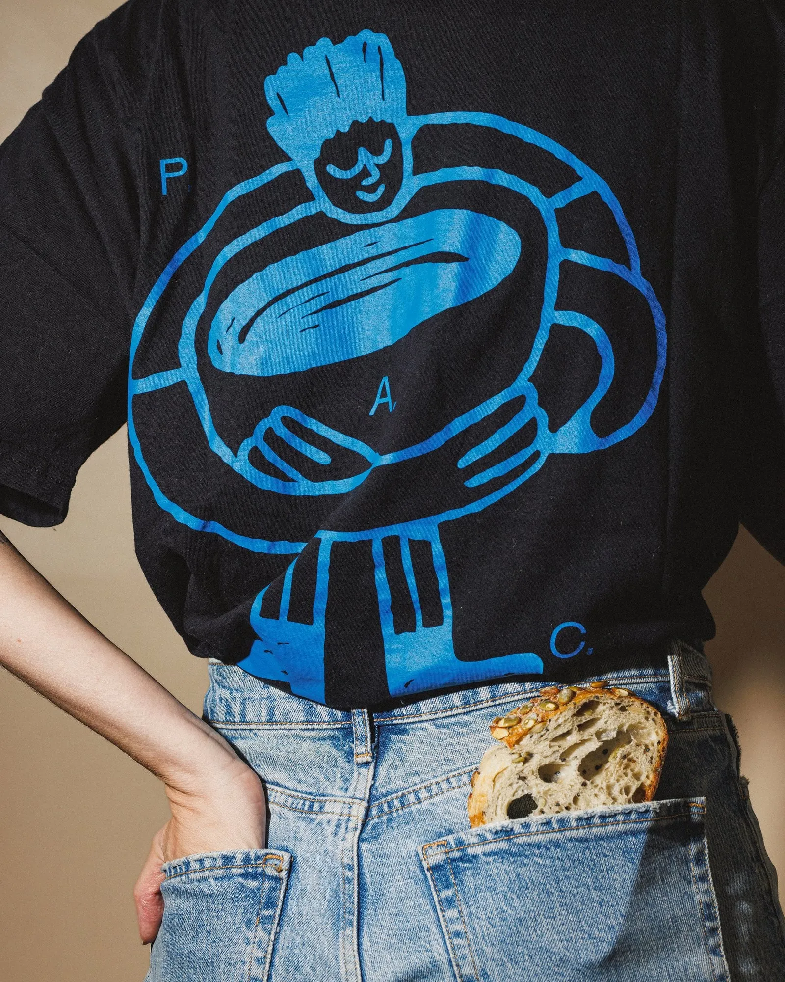 Camiseta negra con una figura azul que abraza un cuenco enorme y las letras «P.A.C.», con una rebanada de pan asomando del bolsillo del pantalón