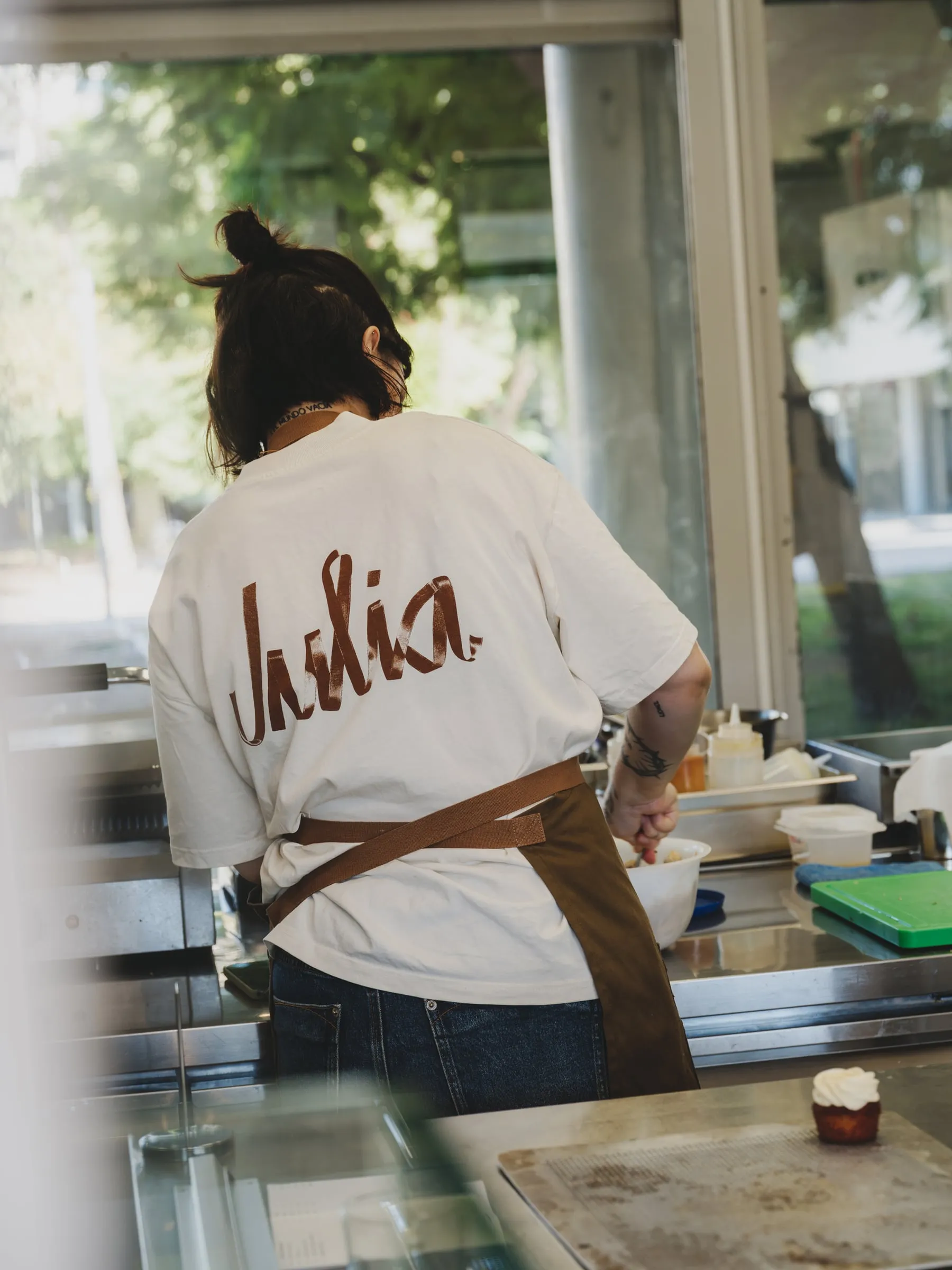 Cocinera de espaldas con camiseta blanca con el logotipo de Julia en marrón y un delantal marrón