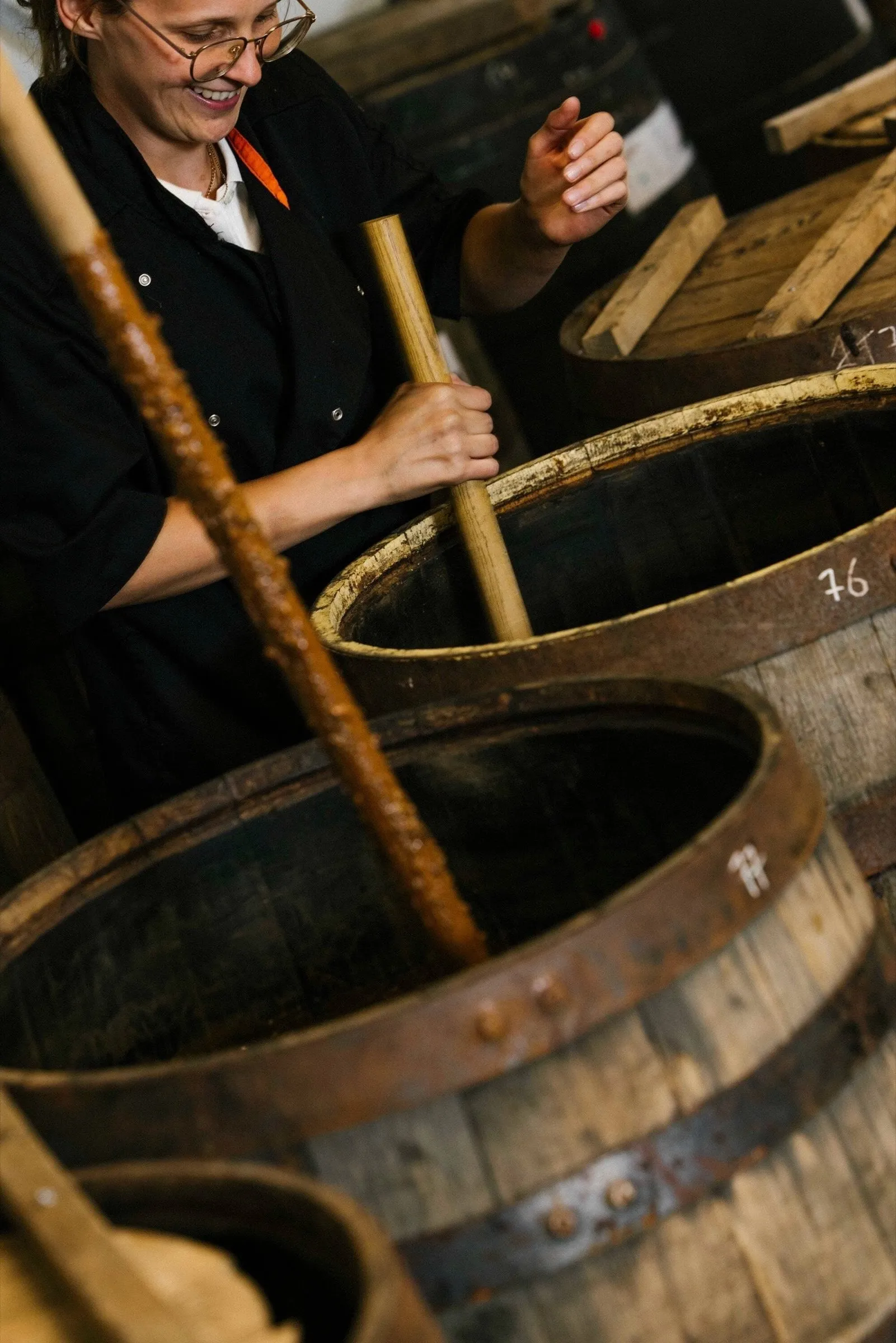 Una trabajadora sonriente remueve con un palo largo una pasta marrón dentro de una barrica de roble con el número 76