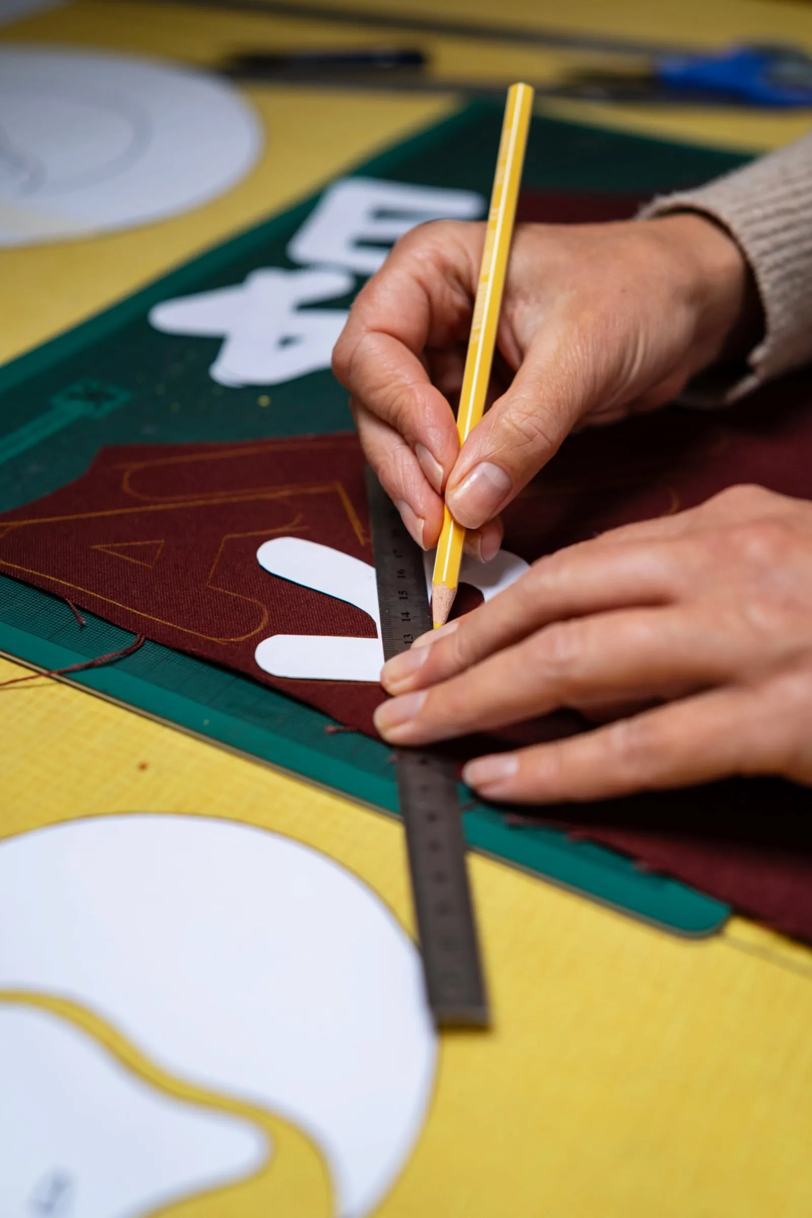 Manos que sostienen un lápiz amarillo y una regla mientras dibujan curvas blancas sobre piezas de tela granate y verde de una bandera
