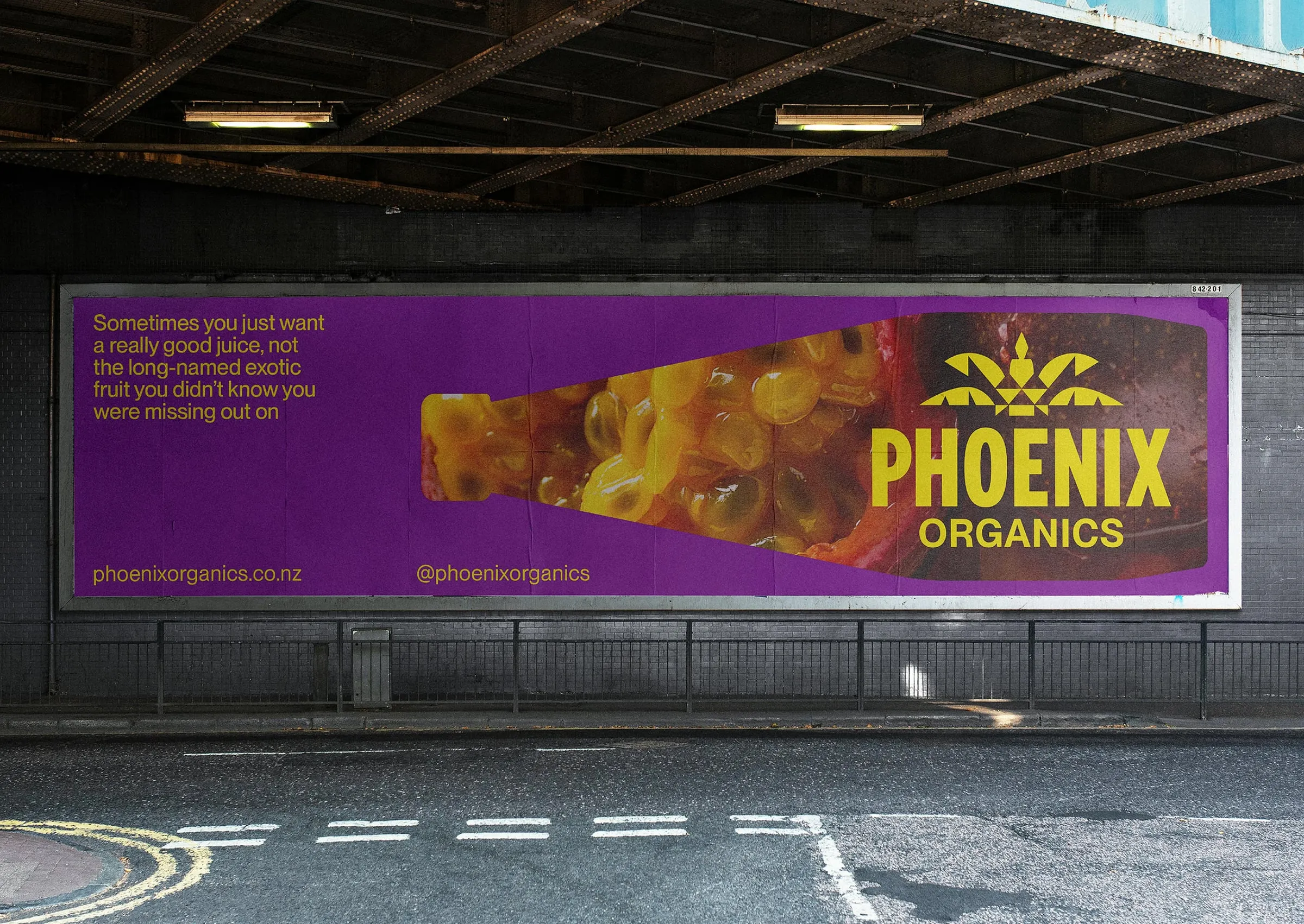 Valla publicitaria morada bajo un puente con la silueta de una botella llena de pulpa de maracuyá y el logotipo de Phoenix Organics en amarillo