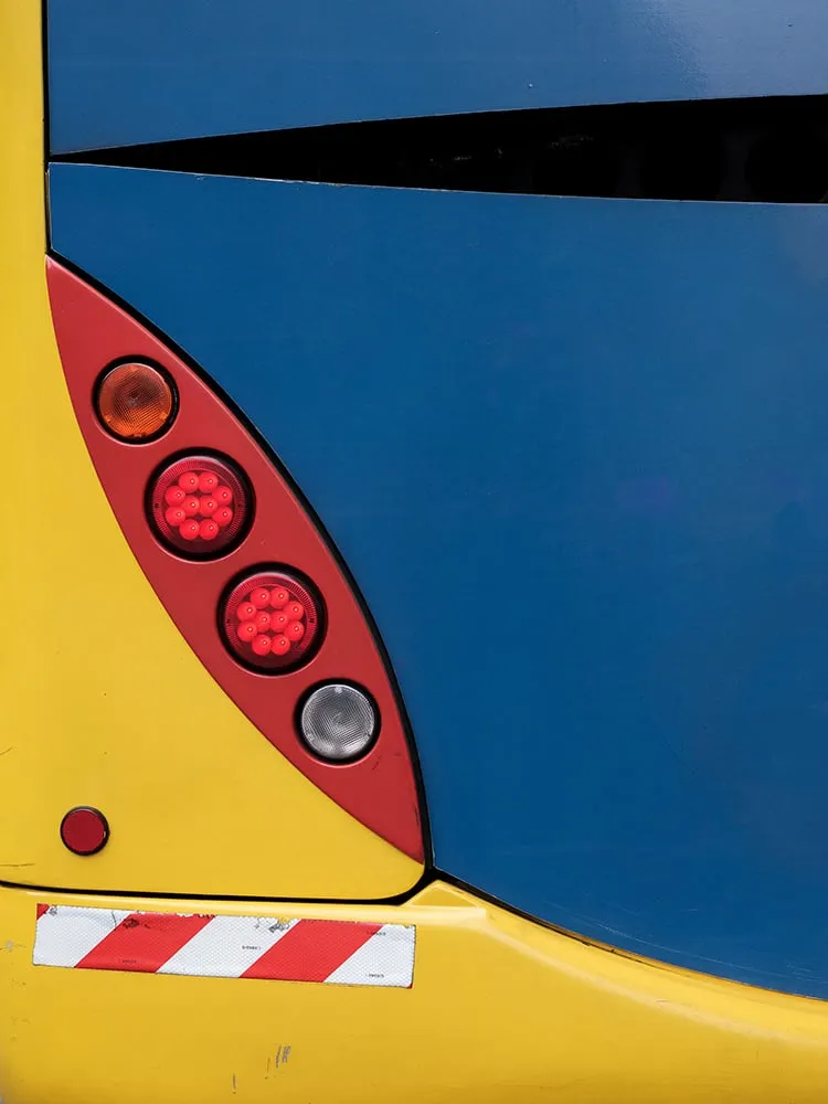 Trasera de autobús en azul oscuro y amarillo con un piloto rojo en forma de gota a la izquierda y una banda de rayas rojas y blancas, como un reflejo de la imagen anterior
