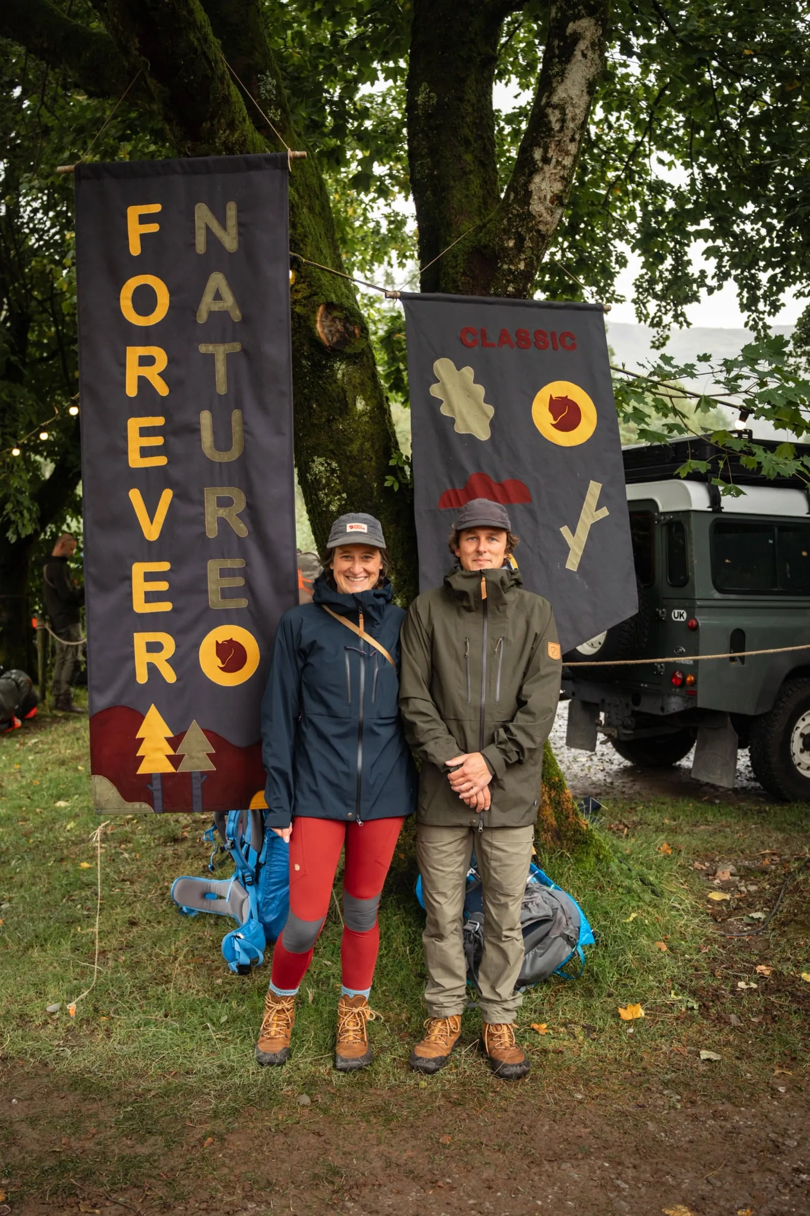 Emma y Andrew Groves, de Miscellaneous Adventures, de pie ante dos banderas de Fjällräven, con un vehículo y árboles cubiertos de musgo detrás
