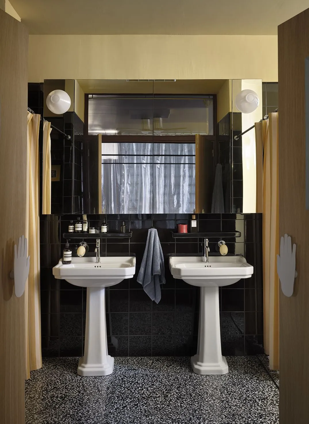 Baño con dos lavabos de pie, azulejo negro, cortinas amarillas y tiradores de puerta en forma de mano