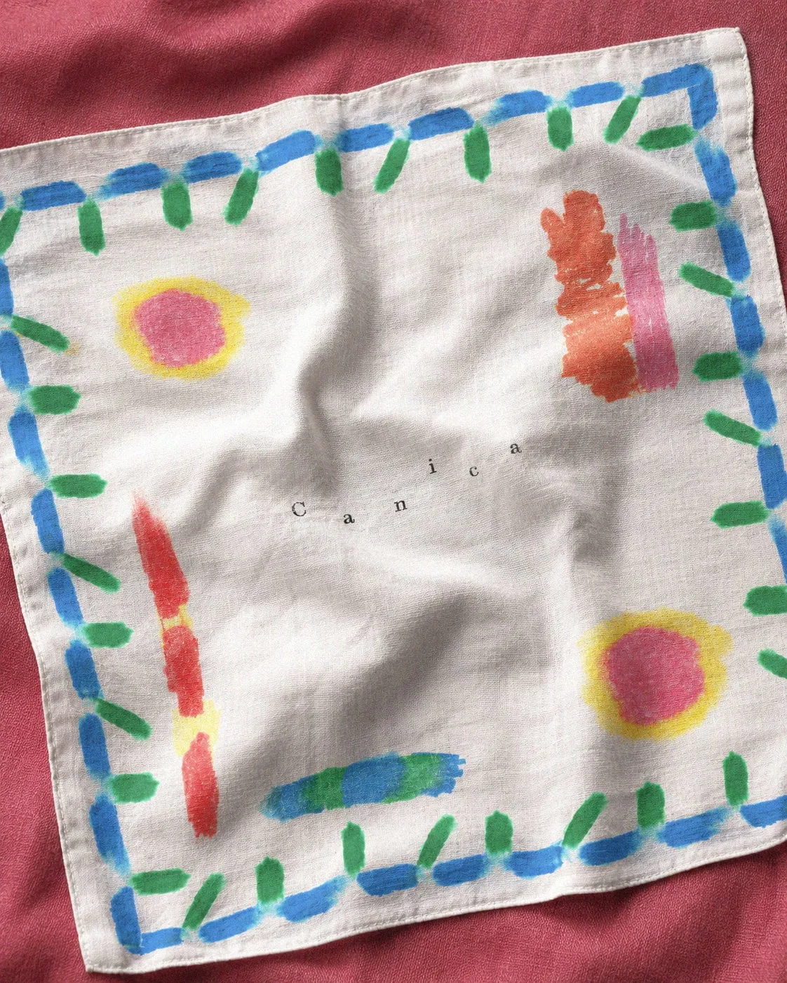 Pañuelo de lino con un borde de trazos azules y verdes y manchas de colores, con la palabra Canica en el centro