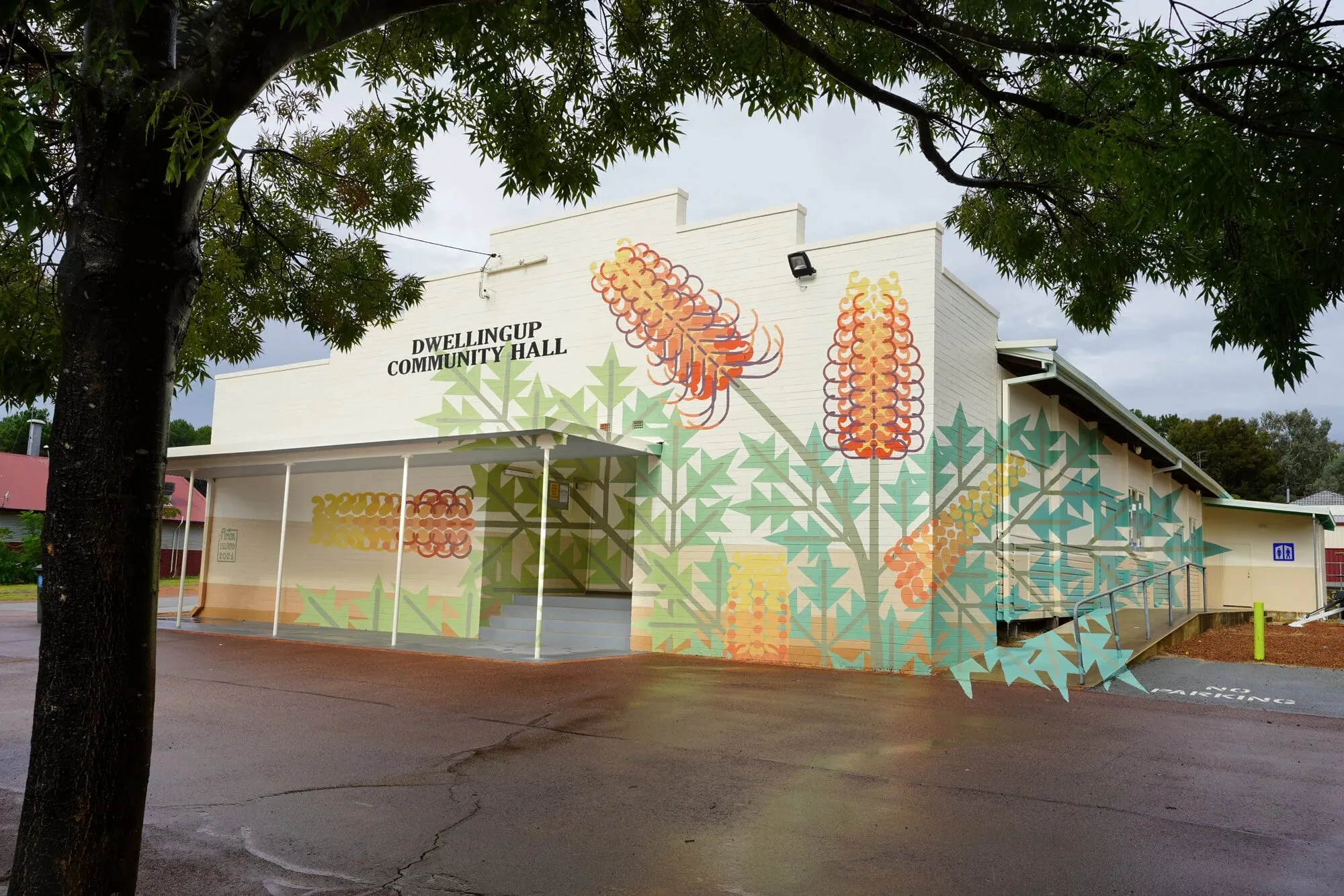 Mural de grevilleas naranjas y hojas sobre la fachada blanca de un salón comunitario en Dwellingup