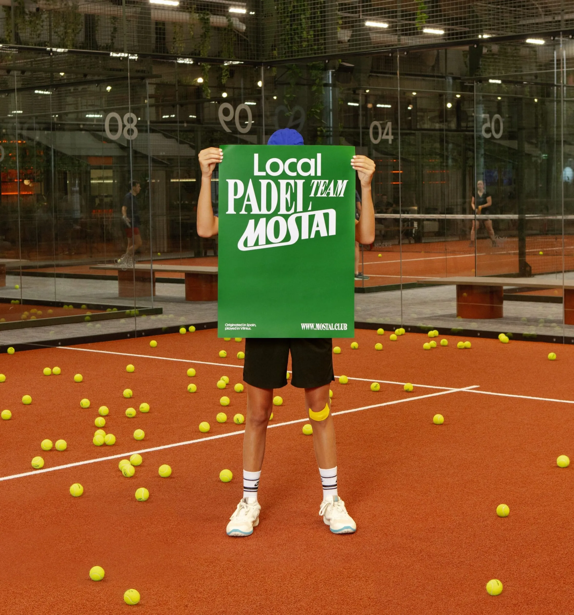 Pala de pádel con manchas negras y una botella verde con el nombre Mostai en vertical, sobre la pista de tierra naranja