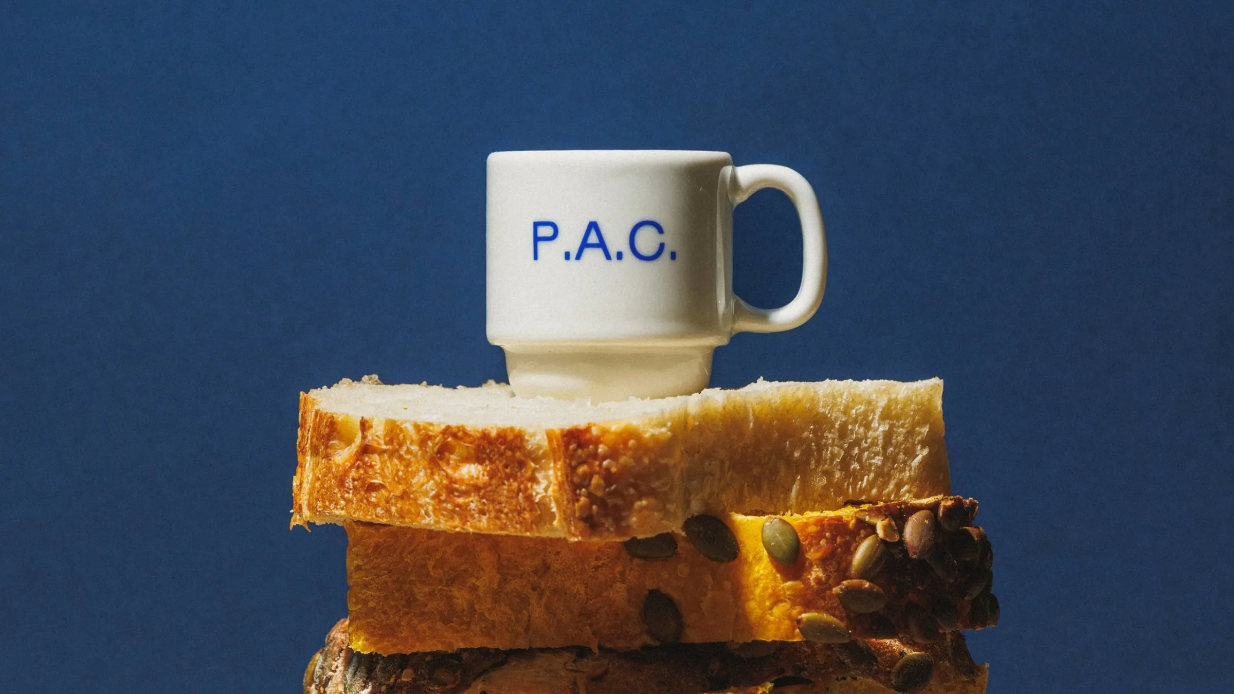 Taza blanca con las letras azules «P.A.C.» apoyada sobre dos rebanadas de pan, con un fondo azul oscuro