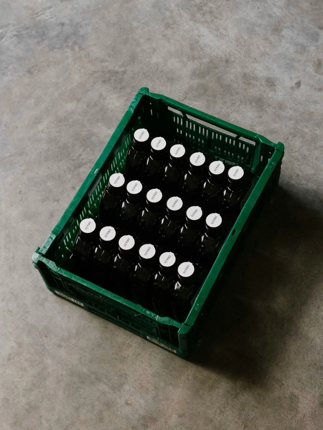 Caja de plástico verde vista desde arriba con dieciocho botellas oscuras de Oraio y tapones blancos sobre un suelo de cemento