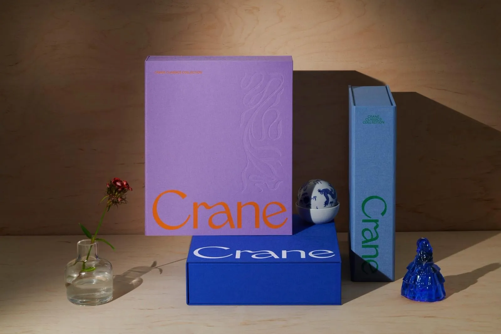 Tres cajas de tela, una violeta con el logotipo naranja, otra azul cobalto y otra azul pizarra, junto a una flor en un jarrón y dos figuras de porcelana y vidrio azules