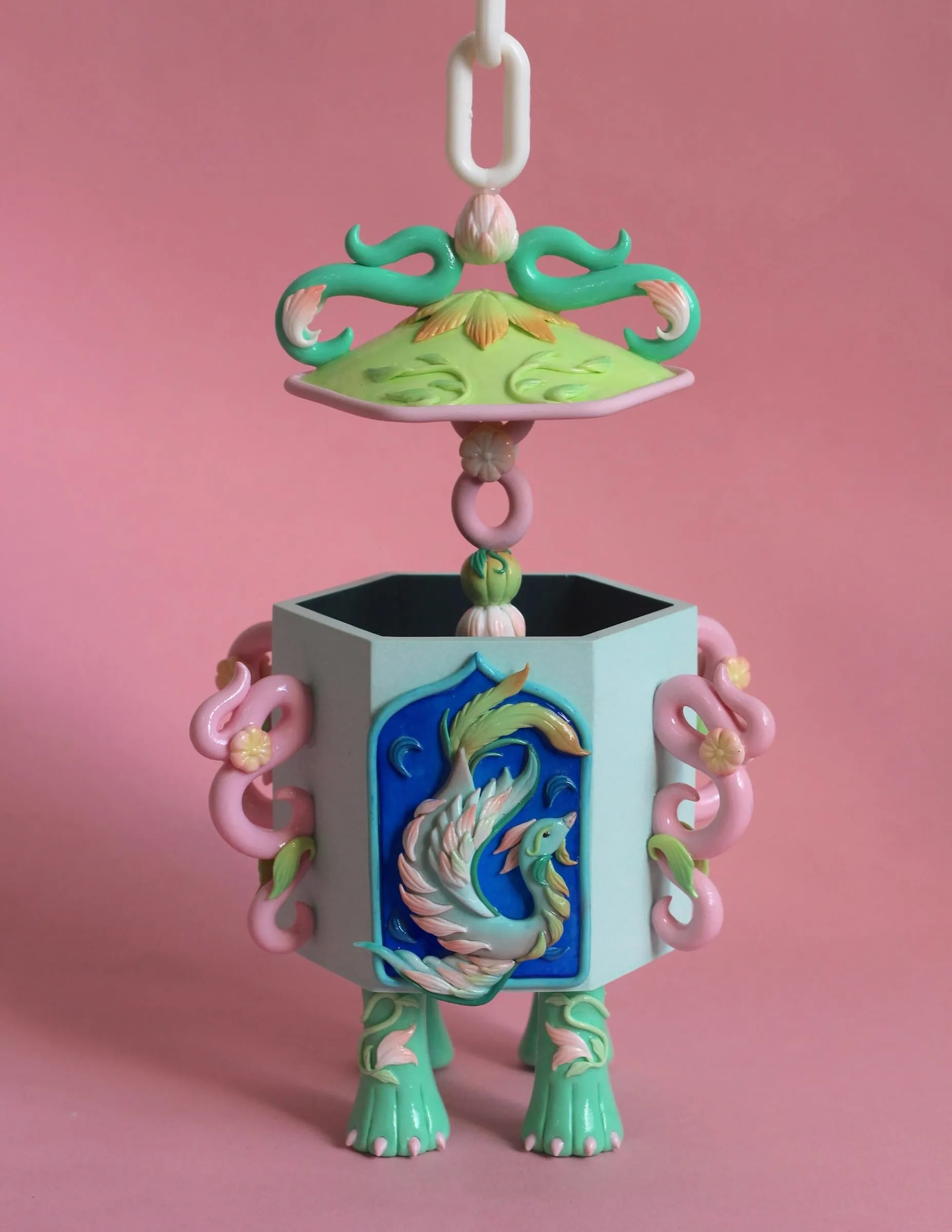 Botella hexagonal azul pálido y rosa con un fénix en un panel azul; la tapa verde cuelga de una cadena blanca