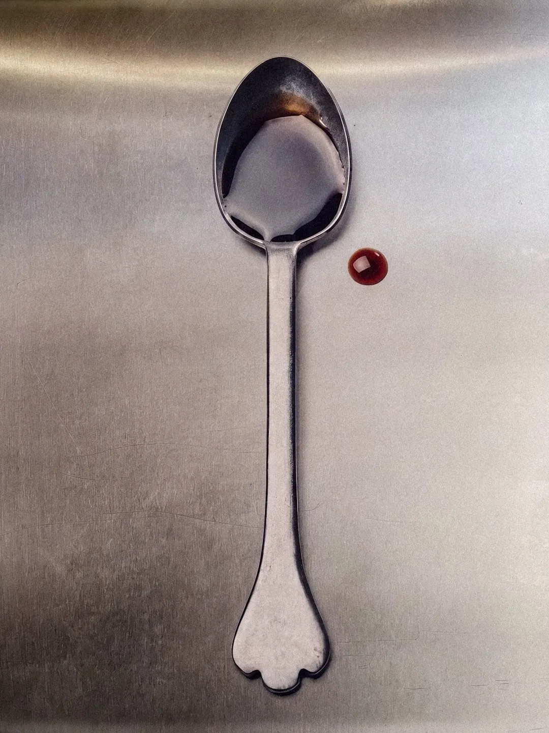 Cuchara de plata sobre una superficie metálica con restos de salsa y una gota roja ámbar al lado