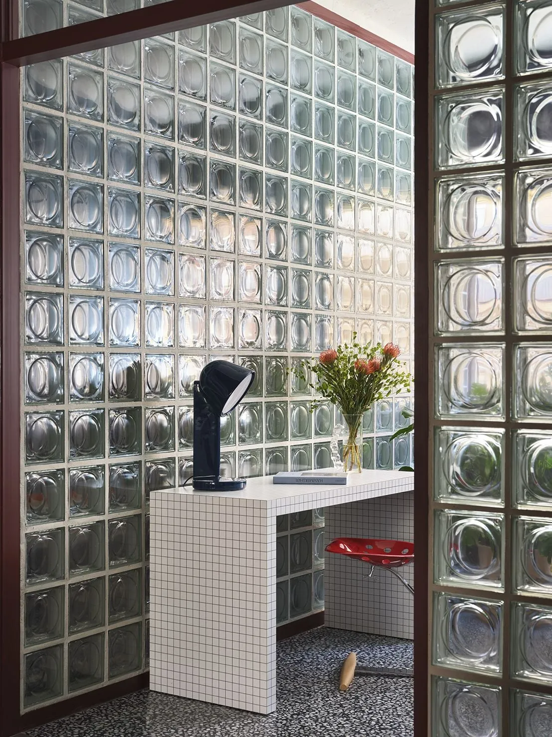 Rincón con una mesa de azulejo blanco cuadriculado, una lámpara de mesa negra y flores, junto a bloques de vidrio