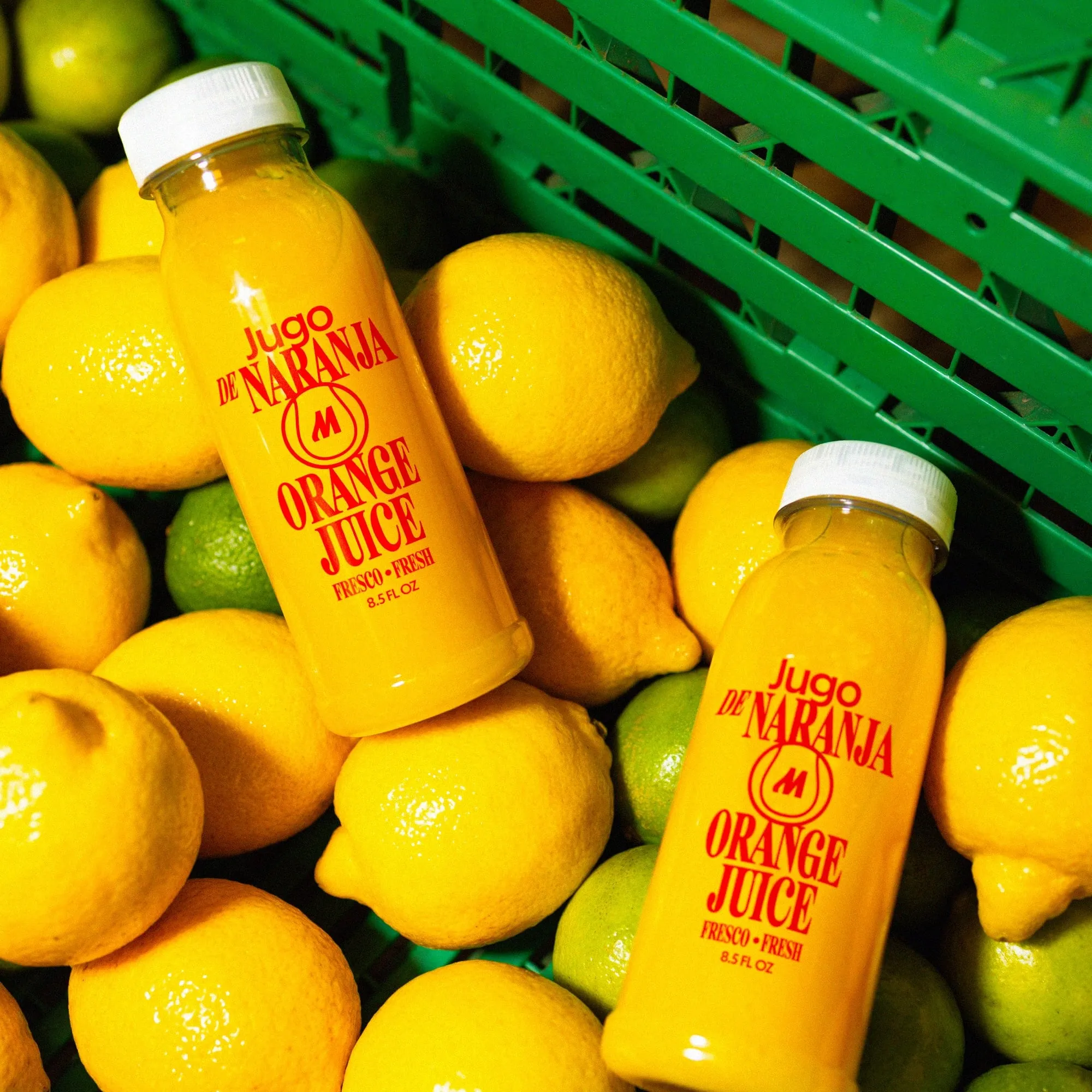 Dos botellas de zumo de naranja con etiqueta roja de Mostai entre limones y una caja verde