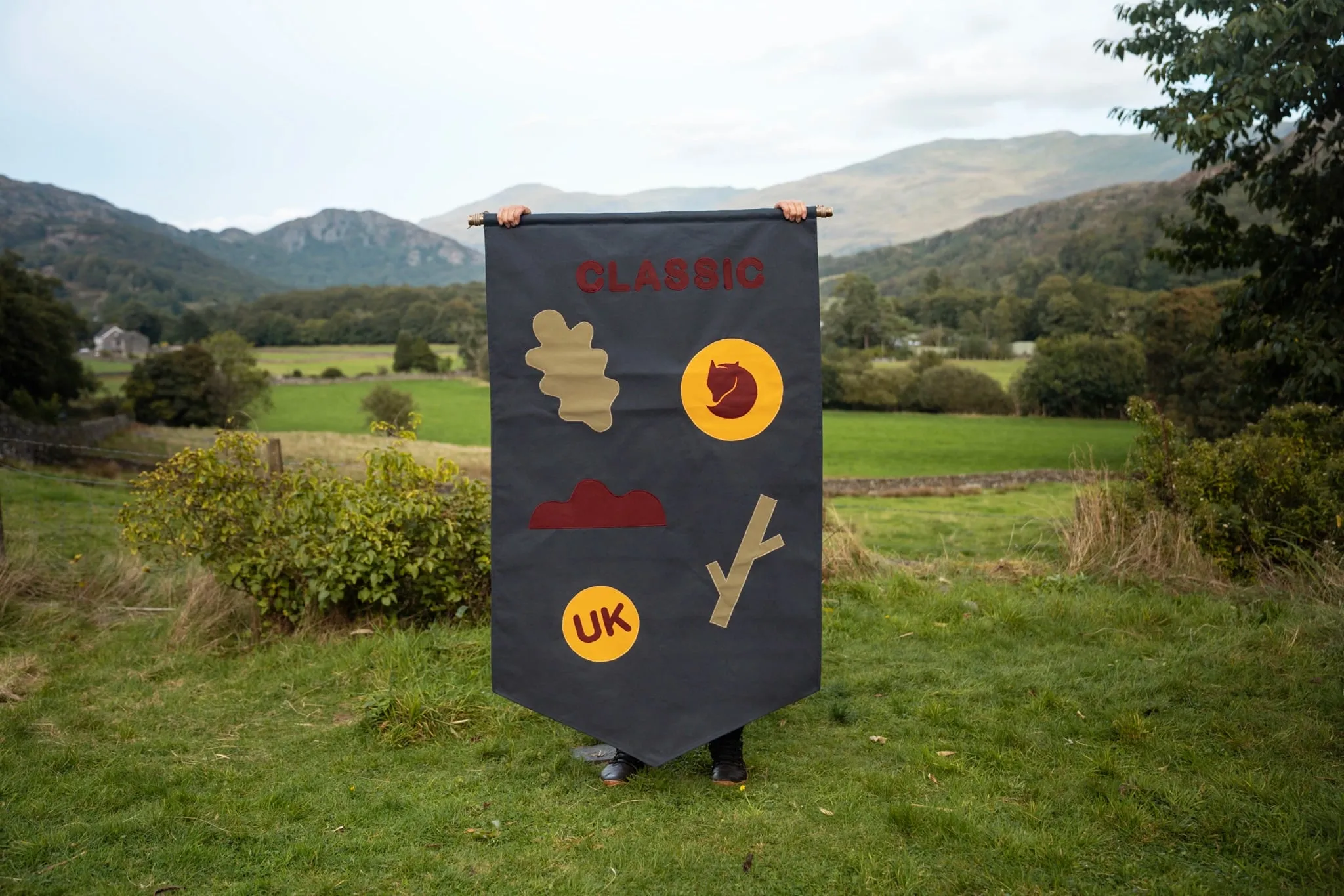 Bandera gris oscura sostenida frente a un valle, con la palabra «Classic» en granate, una hoja de roble, el zorro de Fjällräven sobre un círculo amarillo y un círculo con «UK»