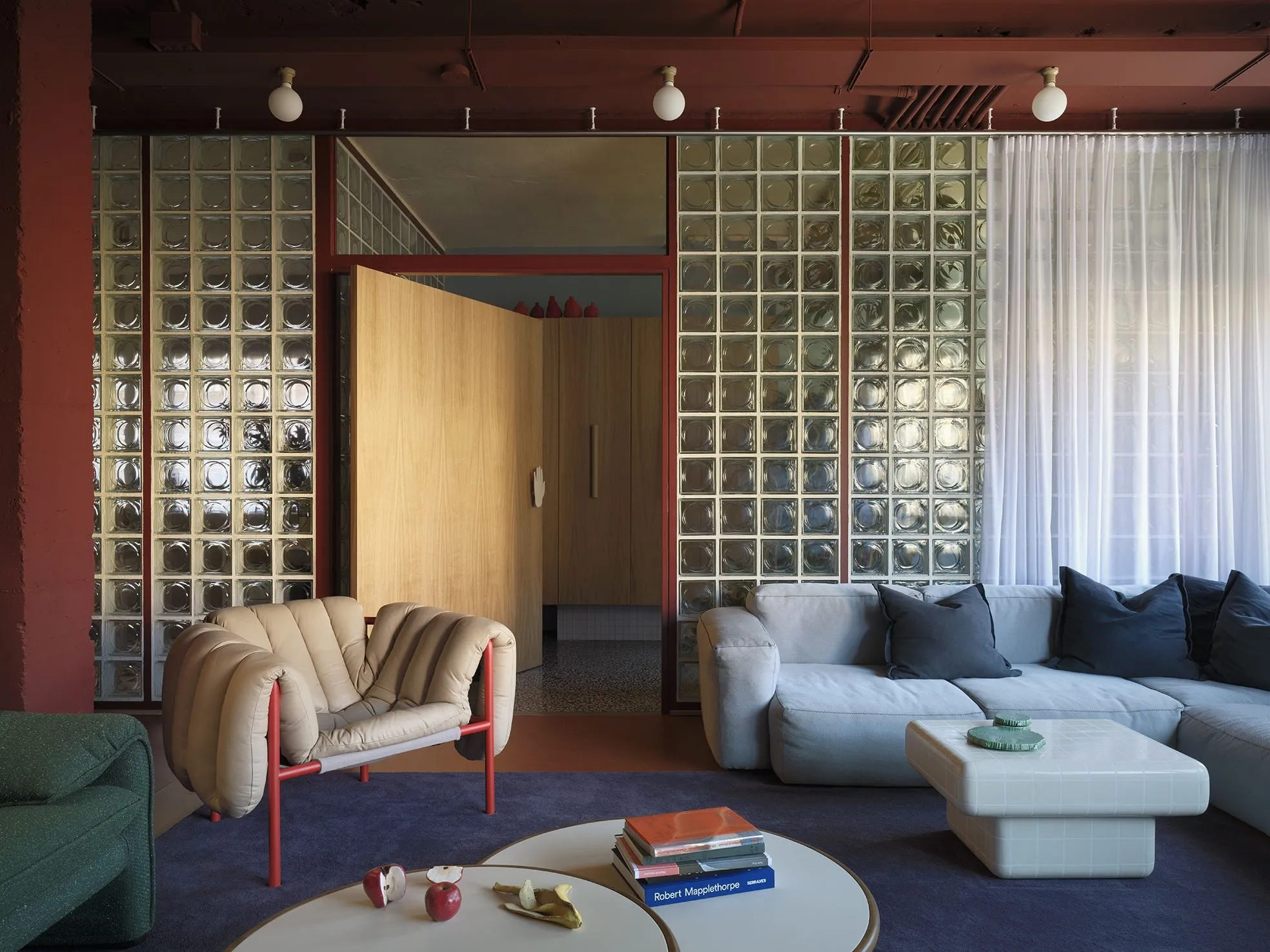 Salón de Villa 2030 en Madrid con un muro de bloques de vidrio, un sofá gris y la butaca beige Puffy de Faye Toogood