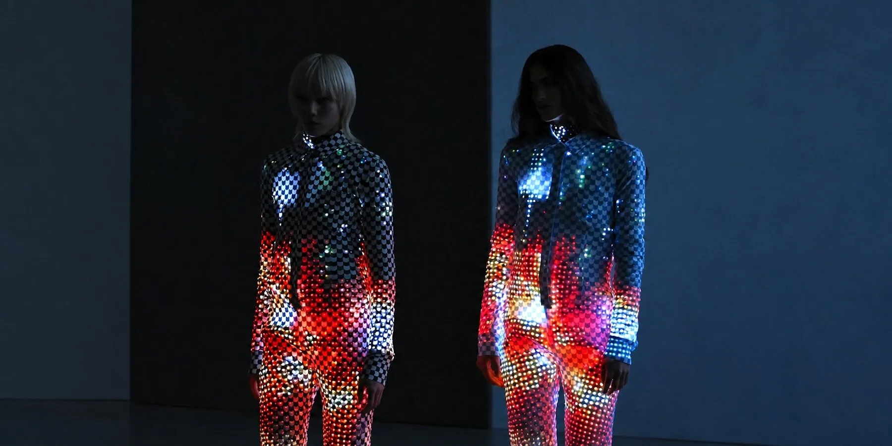Dos modelos de pie en una sala oscura, con trajes de cuadros cubiertos de luces de colores rojas, azules y amarillas desde el torso hasta las piernas