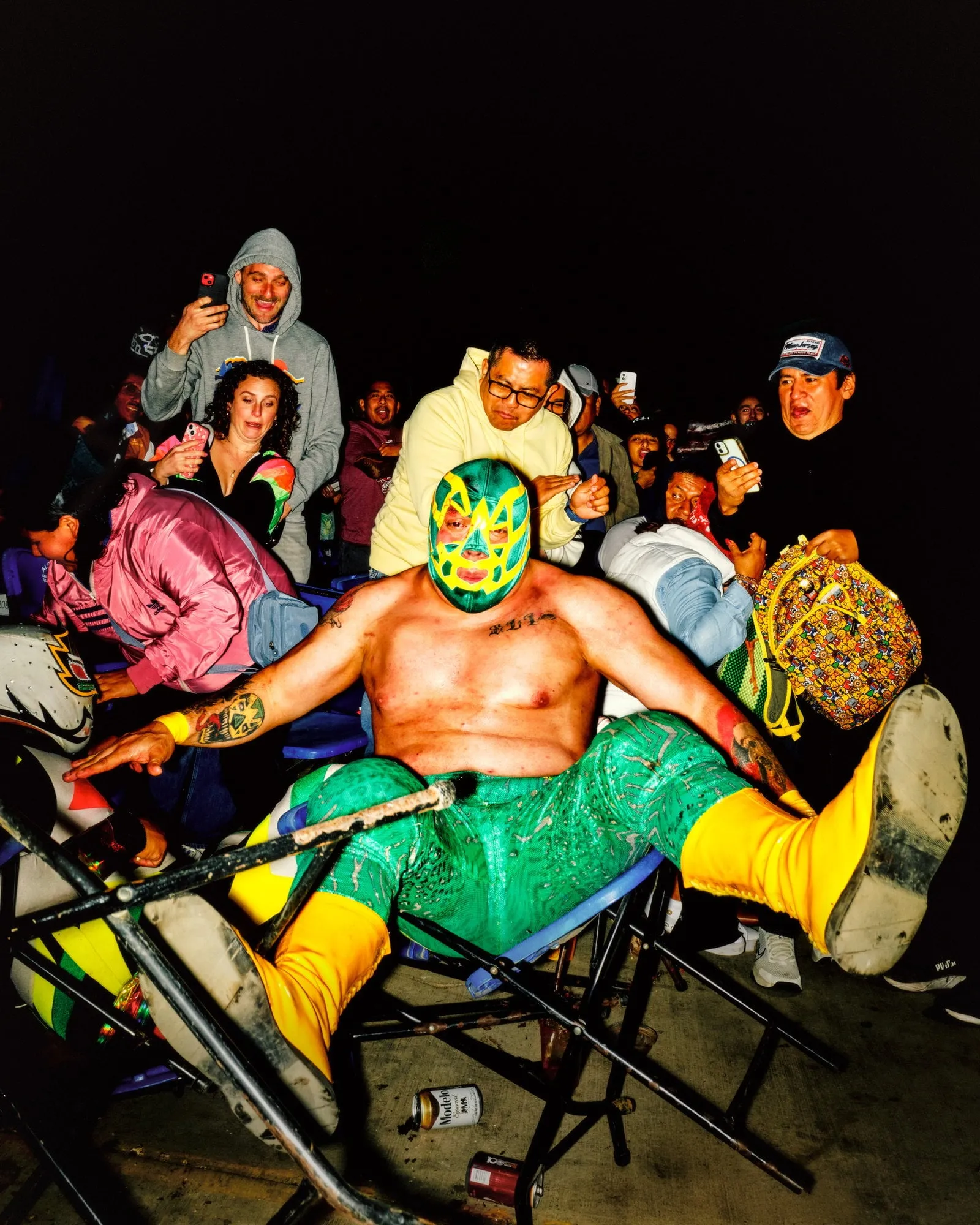 Luchador enmascarado tumbado entre sillas azules numeradas y volcadas en un pasillo del público, con espectadores al fondo