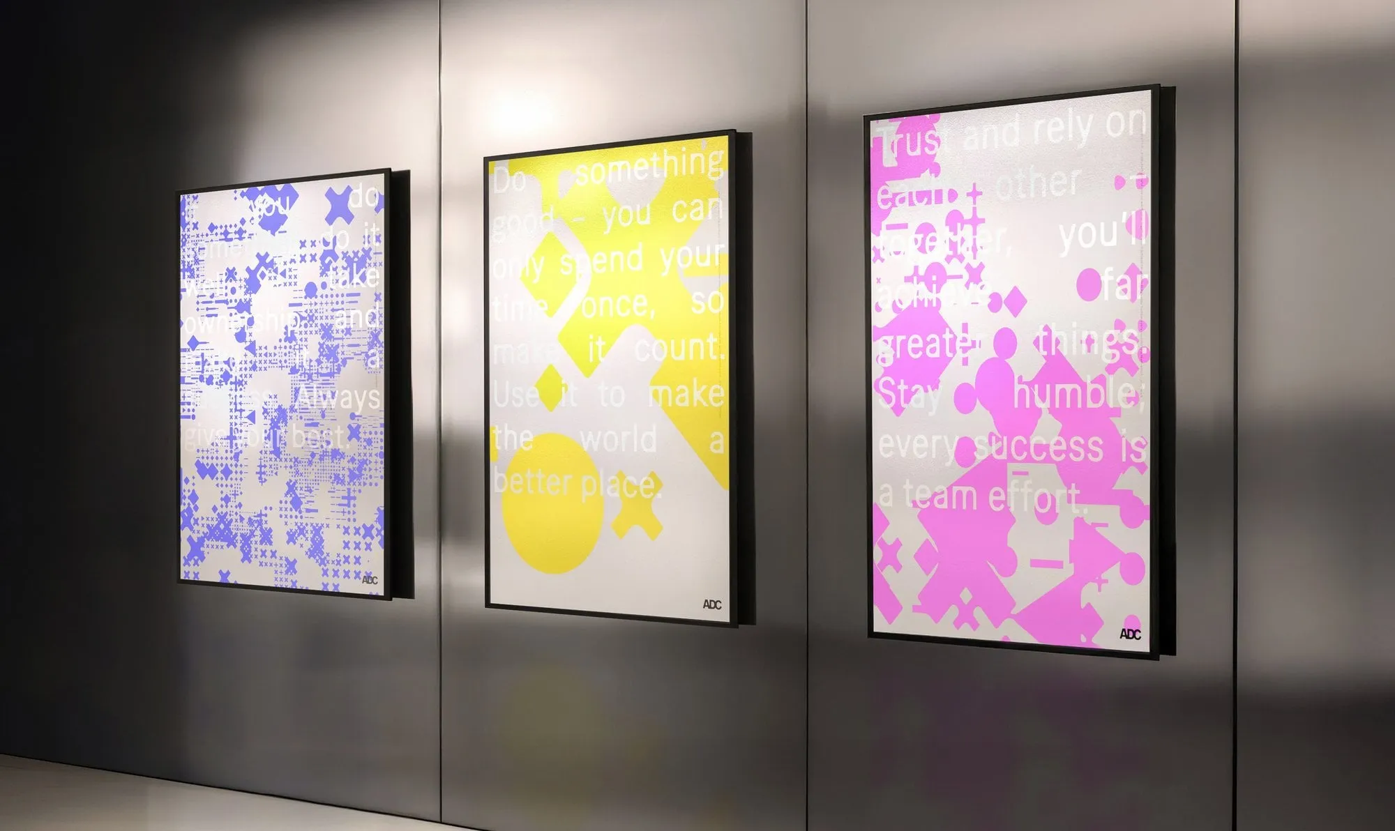Tres carteles enmarcados en una pared metálica con patrones de cruces y rombos en violeta, amarillo y rosa sobre frases en blanco
