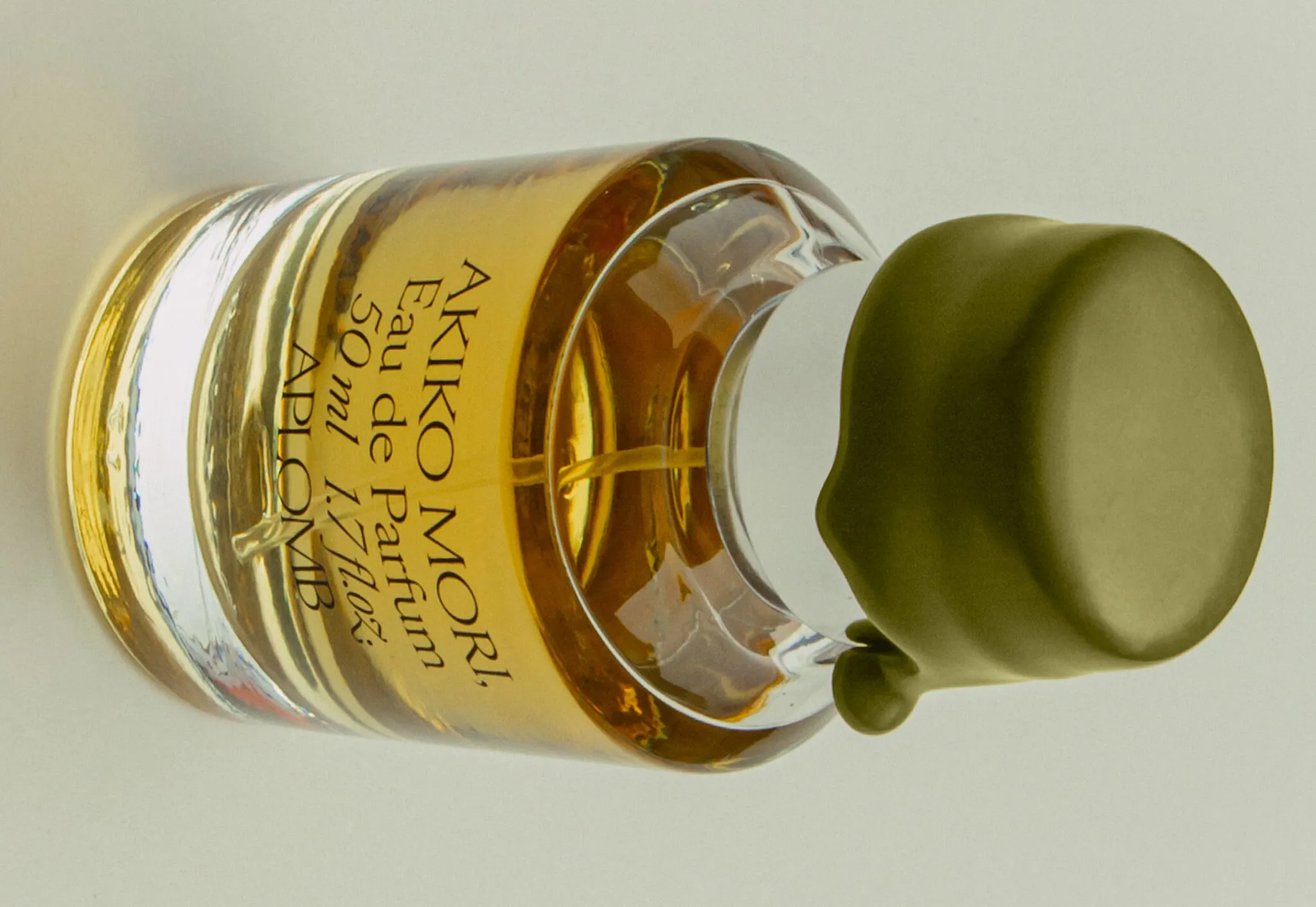 Frasco de perfume APLOMB tumbado, con el líquido ámbar, la etiqueta «Akiko Mori» y una tapa plateada cubierta de cera verde oliva con una gota