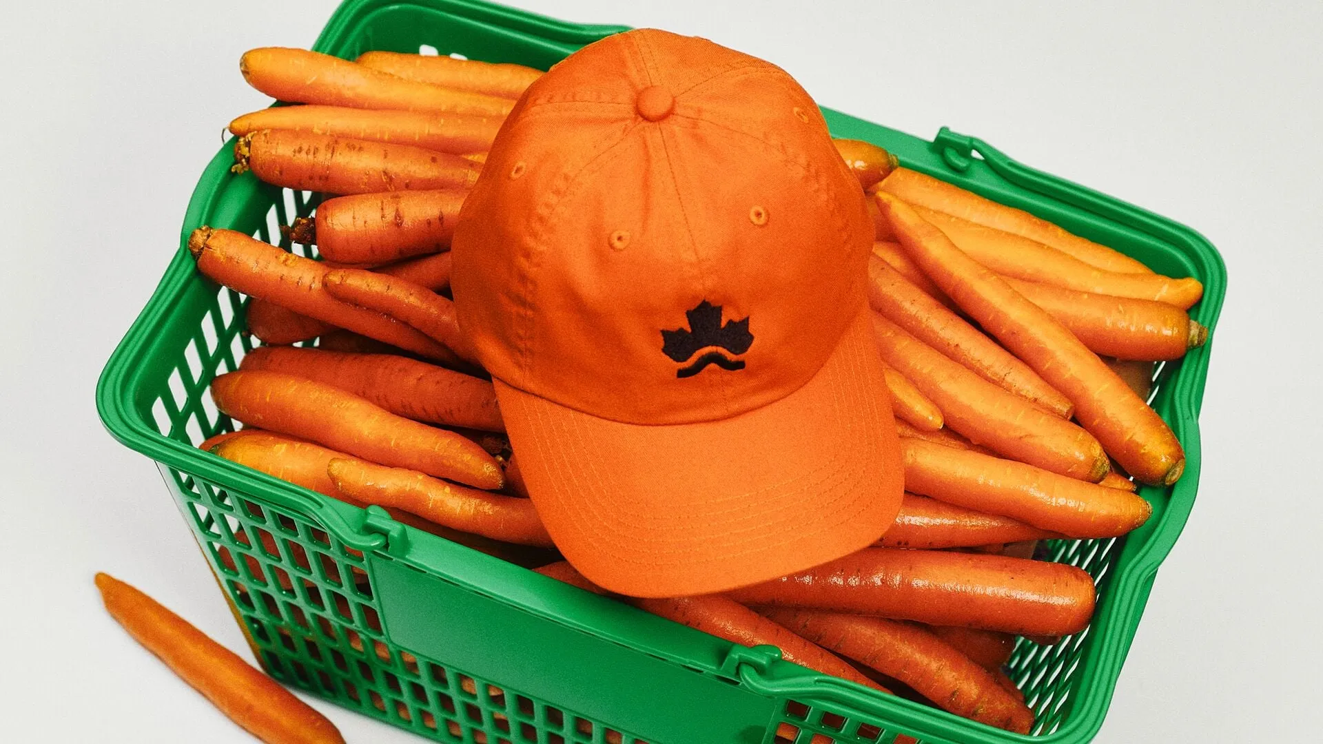 Gorra naranja con una hoja de arce bordada en negro sobre una cesta de plástico verde llena de zanahorias, vista desde arriba