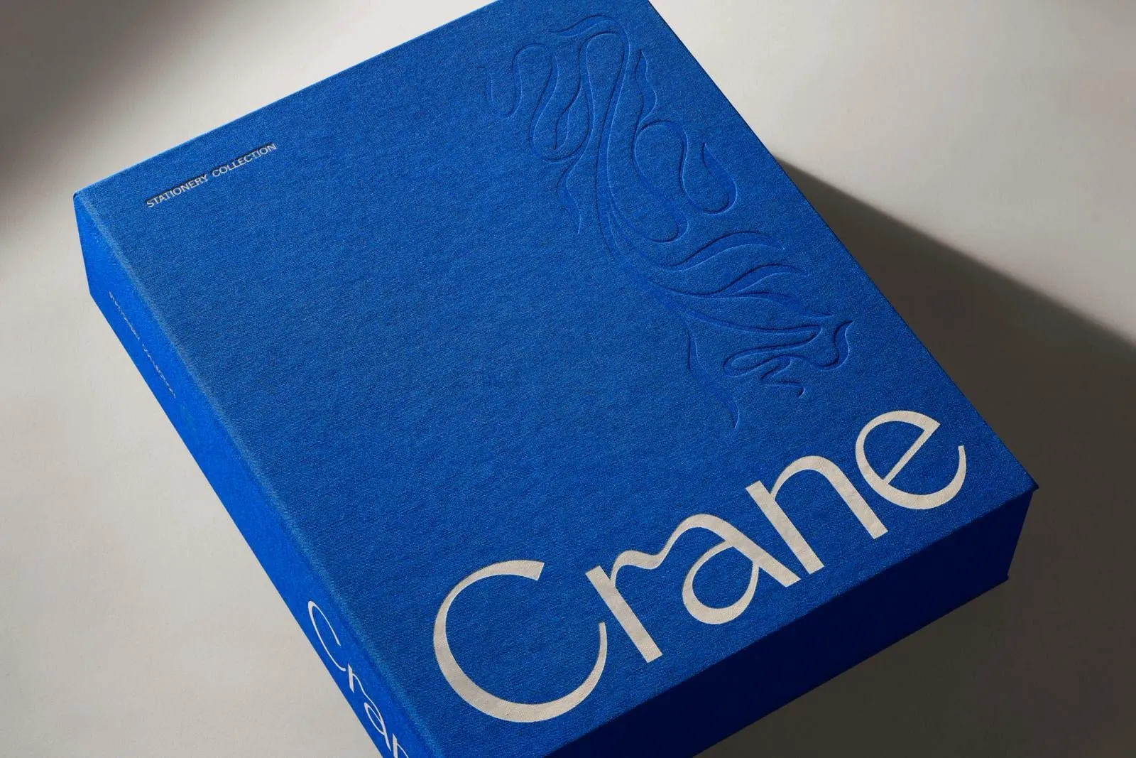 Caja rígida forrada en tela azul cobalto con el logotipo Crane en blanco y un relieve de plantas en la esquina superior, sobre fondo gris claro