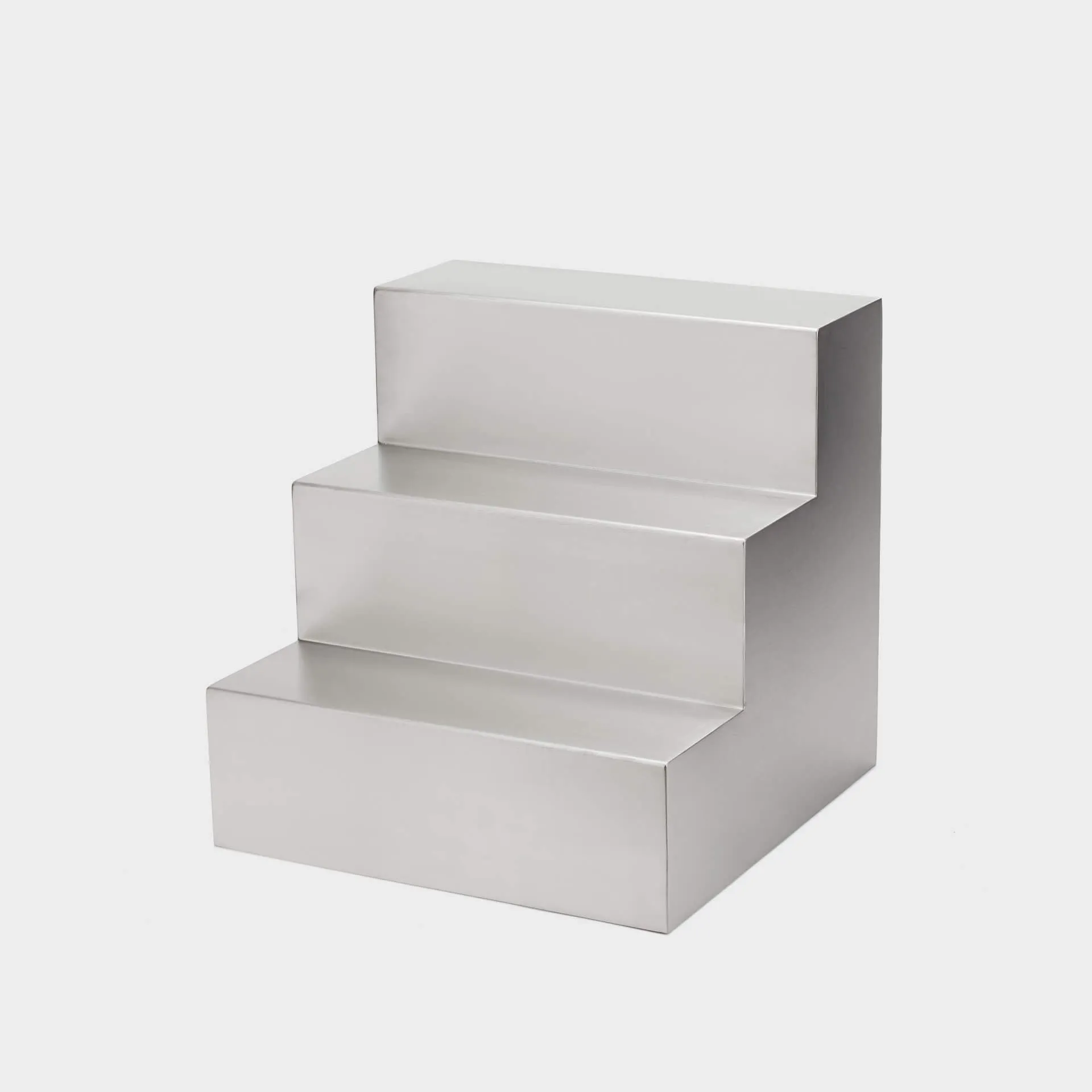 Mesa auxiliar Escala de acero inoxidable con forma de escalera de tres peldaños, vista en perspectiva sobre fondo gris claro