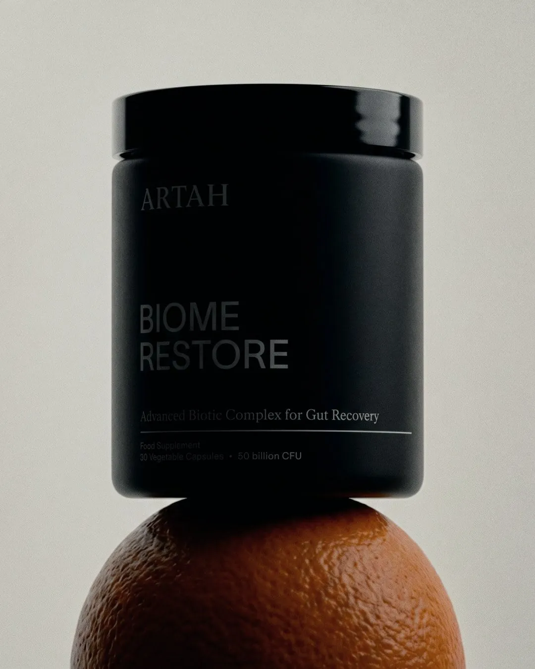 Bote negro mate de Artah con la etiqueta «Biome Restore» en gris, en equilibrio sobre una naranja, con fondo gris claro