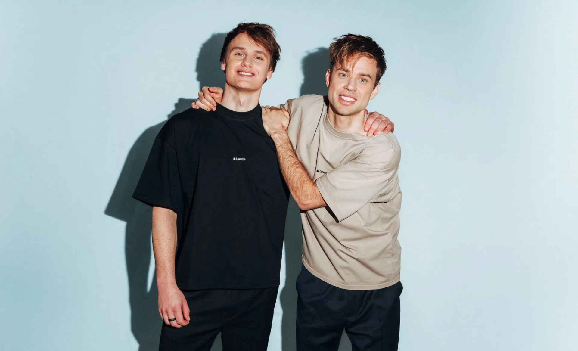 Anton Osika y Fabian Hedin, cofundadores de Lovable, posando abrazados por los hombros con camisetas negra y beige sobre fondo azul claro