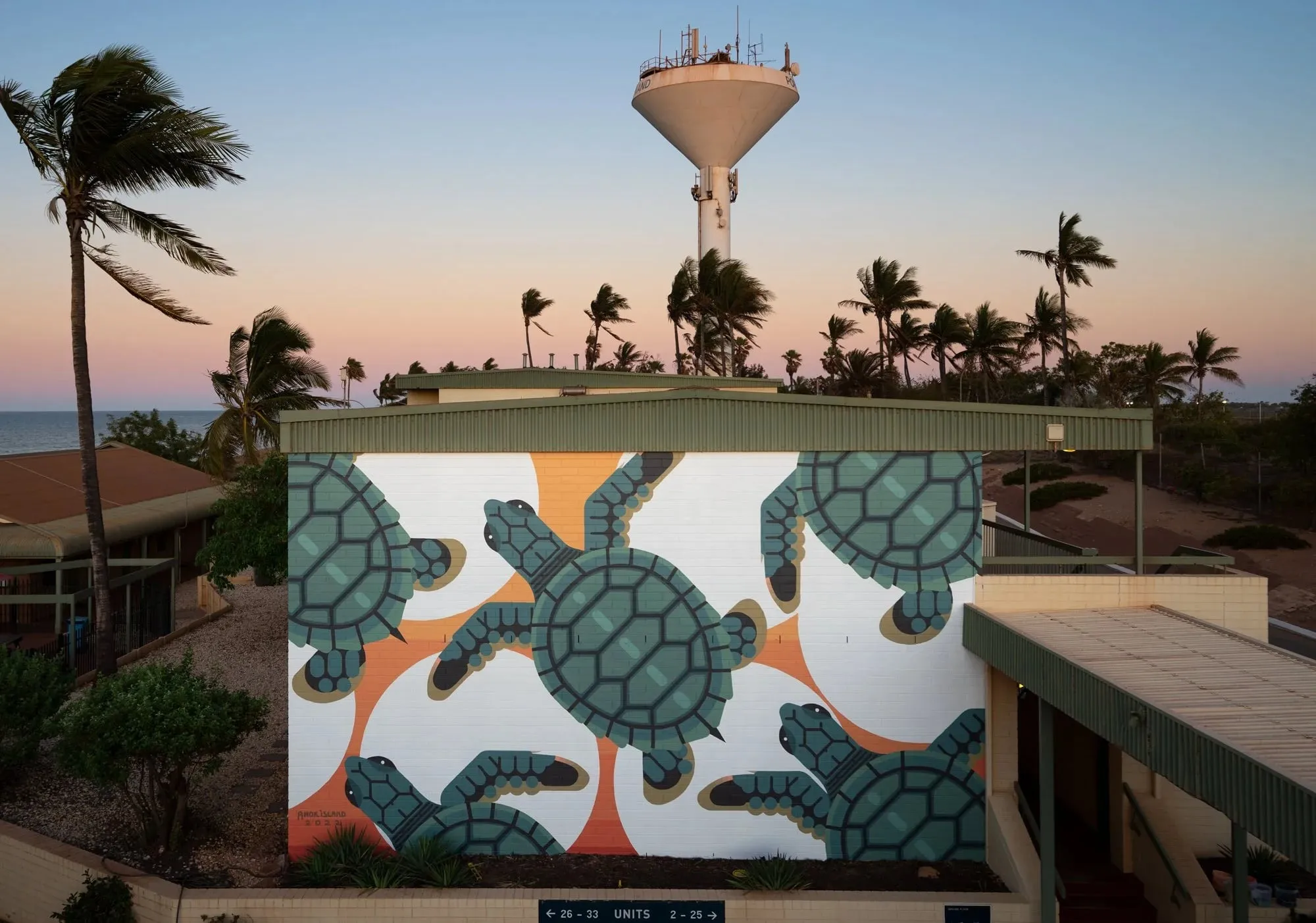 Muro de un edificio bajo un depósito de agua, con cuatro tortugas marinas geométricas en verde y naranja sobre fondo blanco