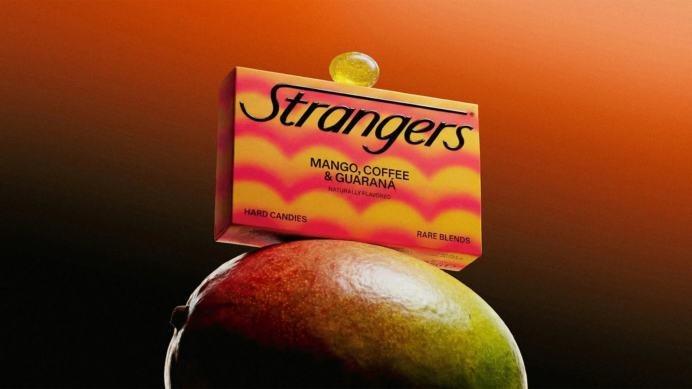 Caja de caramelos Strangers de mango, café y guaraná, con estampado de ondas naranjas y rosas, apoyada sobre un mango maduro con un caramelo encima